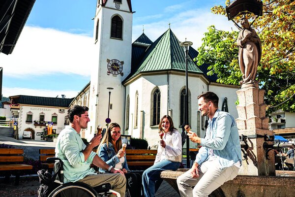 A group of four, including a man in a wheelchair, share laughter and ice cream in a picturesque town square by a church.