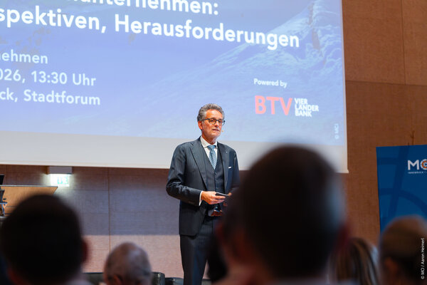 Der Vortragende, mit Brille und grauen Haaren, präsentiert auf einer Konferenz, während das Publikum aufmerksam zuhört.