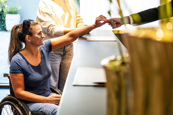 At a hotel front desk, a woman in a wheelchair exchanges a card with a staff member, highlighting accessibility and service.