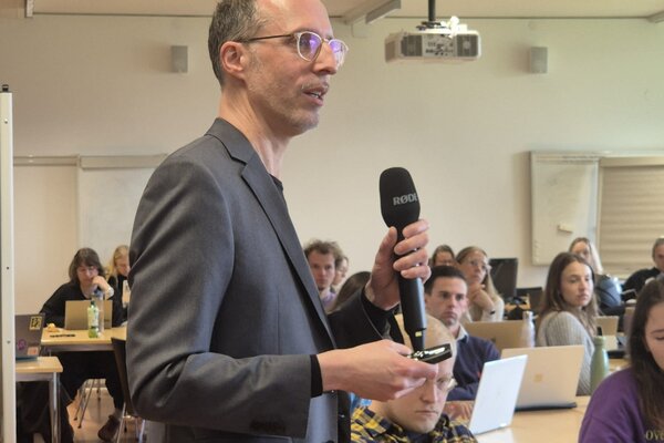 A speaker in a gray suit passionately addresses an audience, holding a microphone while gesturing with his hands.