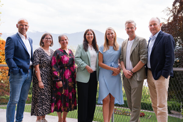 A diverse group of seven individuals stands together outdoors, smiling against a backdrop of mountains and greenery.