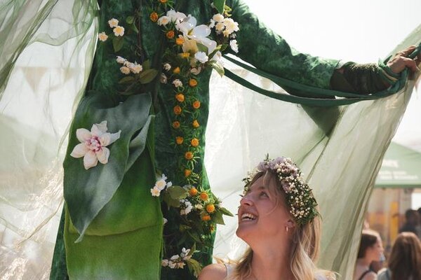 A performer dressed in vibrant green with floral decorations stands tall, delighting a smiling woman in a flower crown.