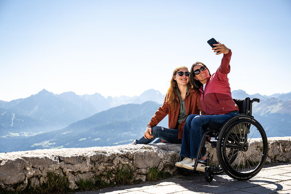 Two women enjoy a sunny day in the mountains, smiling and taking a selfie, with one in a wheelchair and the other beside her.