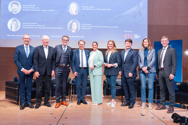 Eine Gruppe von Geschäftsleuten steht auf einer Bühne, umgeben von Stühlen und einem großen Bildschirm, der Informationen über die Veranstaltung zeigt.