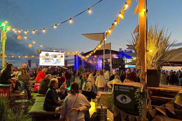 A lively outdoor event scene filled with people enjoying food and drinks under colorful lights and decorations at dusk.