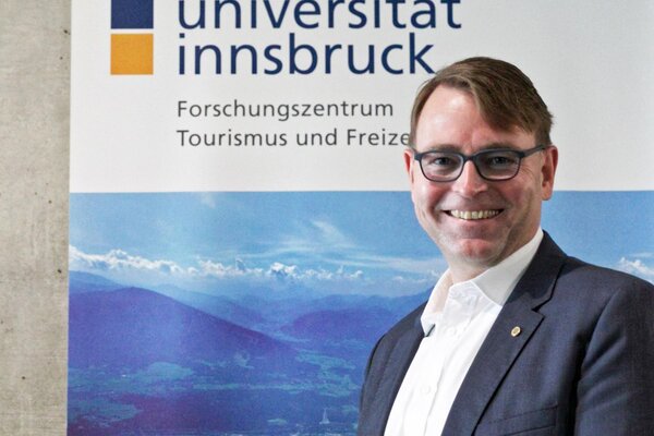 Der Mann in Anzug und Brille präsentiert sich vor einem Hintergrund mit dem Logo der Universität Innsbruck und Berglandschaft.