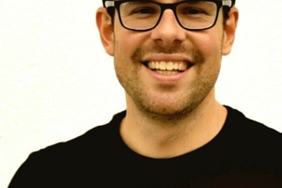 Der Mann mit Brille hat kurzes, dunkles Haar und zeigt ein freundliches Lächeln in einem neutralen Hintergrund.