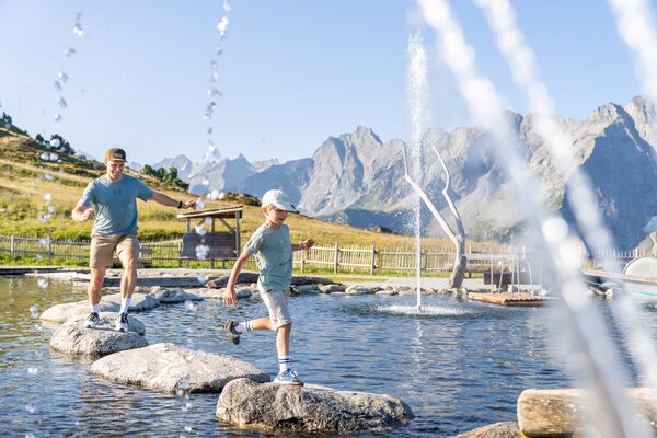 Mit einem Lächeln auf dem Gesicht springt ein Kind über große Steine in einem Wasserspiel, umgeben von beeindruckenden Bergen.