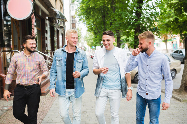 Casually dressed, four men share a lighthearted moment, walking together on a sunny street filled with trees and shops.