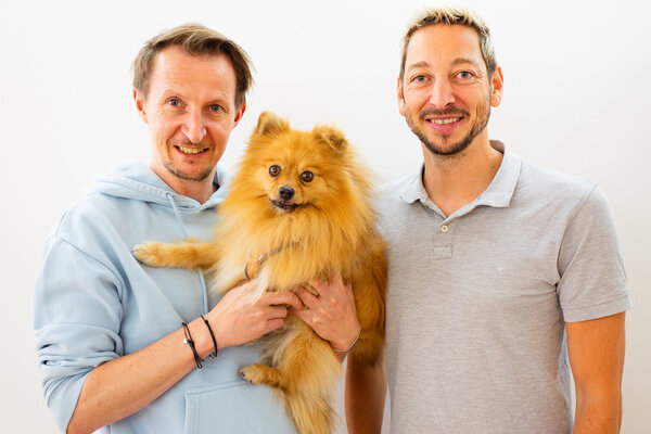 Two smiling men stand side by side, one holding a fluffy Pomeranian dog against a bright, neutral background.
