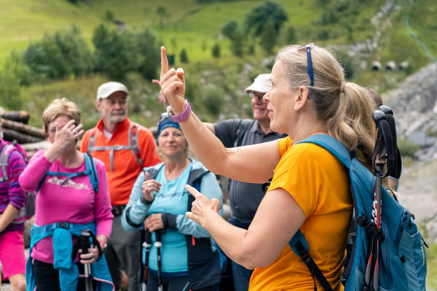 Sechs Wanderer stehen im Freien, während eine Frau ihnen mit ausdrucksvoller Mimik und Handzeichen etwas vermittelt.