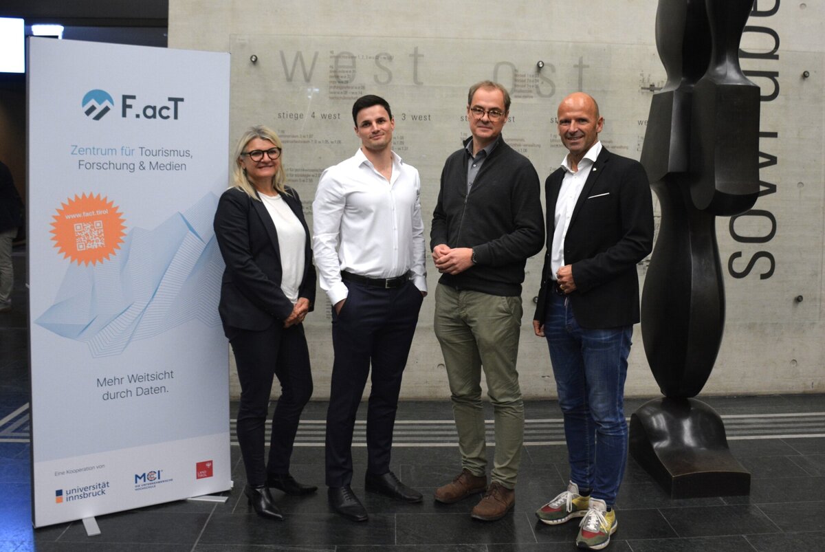 Im Hintergrund ist ein großes Informationsschild sichtbar, das Details über das F.actT-Zentrum enthält, während die Personen freundlich wirken.