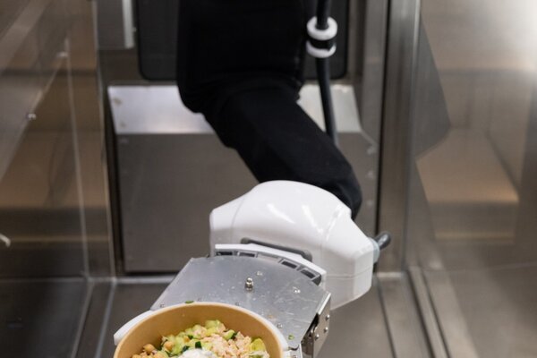 Ein Roboterarm reicht eine Schüssel mit frischem Salat, garniert mit Joghurt und Oliven, in einem modernen Essbereich.