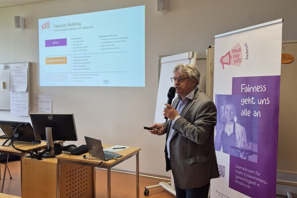 A speaker presents on capacity building, holding a microphone, with a projector displaying key points in a conference room.