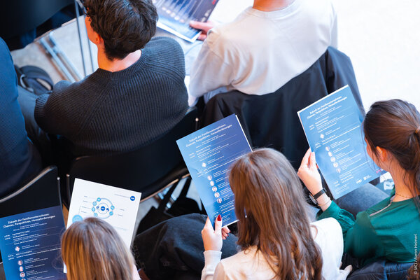 Eine Gruppe von Menschen sitzt in einem Raum und hält Programmhefte in den Händen, während sie auf eine Veranstaltung warten.